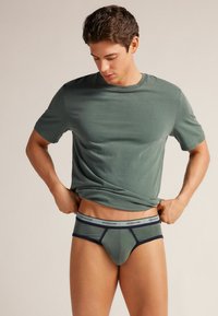 Grünes Baumwoll-T-Shirt, kombiniert mit passenden grünen Shorts mit einem marineblauen Bund. Die Passform ist eng und betont die Körperform des Trägers.