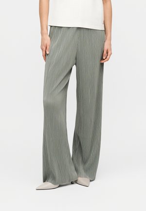 Žena má na sebe voľné, plisované svetlosivé nohavice v páse a špicaté béžové topánky, stojí pred jednoduchým bielym pozadím.