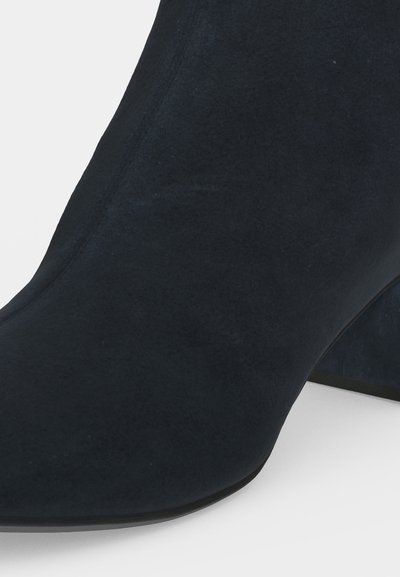 Gros plan d'une bottine en daim noir avec un bout pointu et un talon bloc sur un fond uni.