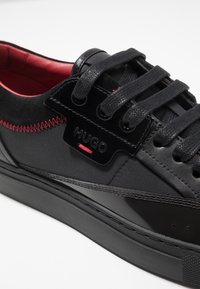 Czarne sneakersy wykonane z teksturowanej tkaniny z błyszczącymi akcentami, z czerwonym wykończeniem, teksturowanymi czarnymi sznurówkami i naszywką z logo po boku.