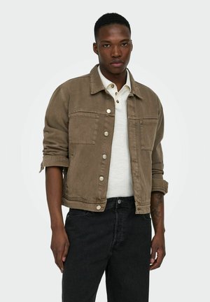 Mężczyzna w brązowej, zapinanej na guziki kurtce dżinsowej, nałożonej na białą koszulę z podwiniętymi rękawami oraz czarnych dżinsach, stojący na jednolitym tle.