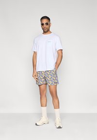 Λευκό βαμβακερό μπλουζάκι με τσέπη, μπλε σορτς με κίτρινο σχέδιο, λευκές κάλτσες εθελοντή και μπεζ αθλητικά παπούτσια. Γκρίζο φόντο.