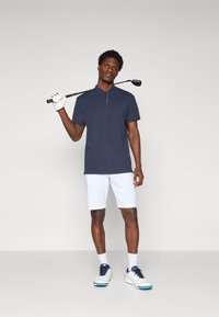 Polo navy cu mânecă scurtă, cu plachetă cu jumătate de nasture, combinat cu pantaloni scurți albi și pantofi de golf albi cu accente albastre, ținând un câmp de golf.