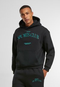 Svart hettegenser laget av myk stoff med "ANOTHER SPORTS CLUB" i turkise bokstaver. Har en frontlomme og ribbet kant.