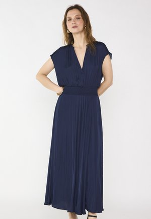 Frau mit welligem Haar, die goldene Ohrringe und ein marineblaues Maxikleid mit kurzen Ärmeln und einem breiten, gerafften Taillengürtel trägt, steht mit den Händen in der Hüfte.