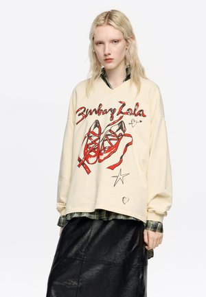 Kremowy, oversize'owy sweatshirt z graficznym nadrukiem czerwonych butów i napisem. Zestawiony z ciemną skórzaną spódnicą i warstwą koszuli w kratę.