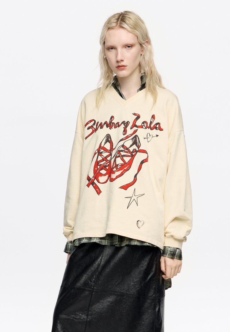 Krémová oversized mikina so grafickou potlačou červených topánok a textu. V kombinácii s tmavou koženou sukňou a vrstvami kockovanej košele.