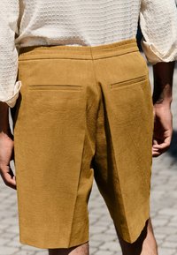 Gulbrune linnedshorts med en afslappet pasform, der har to baglommer og en subtil tekstur, iført en let, tekstureret hvid skjorte.