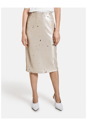 Femme portant une jupe beige à paillettes longueur genou, des talons blancs pointus et un haut blanc, debout, les bras détendus le long du corps.