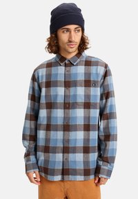 Camicia in flanella con un motivo a quadri blu e marrone, maniche lunghe, chiusura con bottoni frontale e taschino sul lato sinistro del petto. Berretto blu navy a coste.