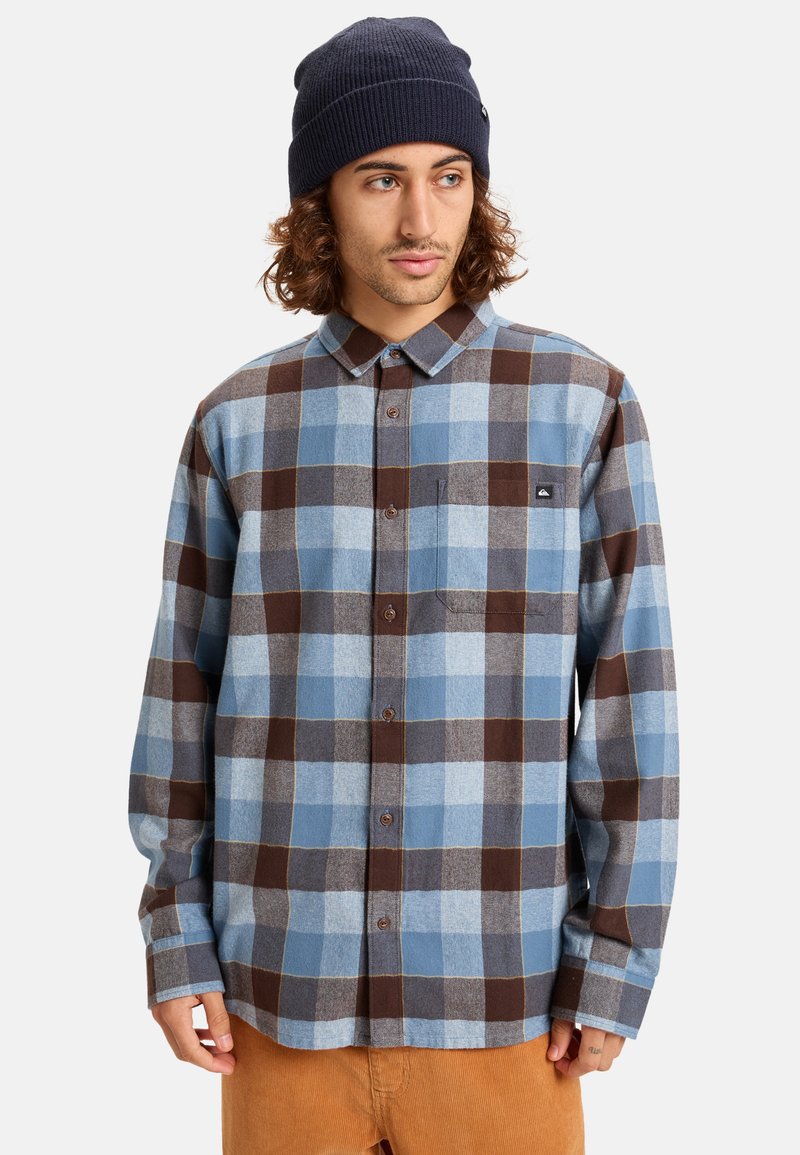 Camicia in flanella con un motivo a quadri blu e marrone, maniche lunghe, chiusura con bottoni frontale e taschino sul lato sinistro del petto. Berretto blu navy a coste.