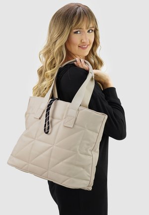 Woman with long blonde hair wearing black holds large beige quilted tote bag over her shoulder against a plain background.
