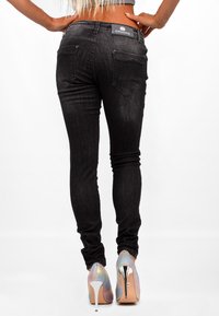Jeans neri attillati con una texture liscia, dotati di due tasche posteriori e di un'etichetta con logo in vita, abbinati a scarpe con tacco alto iridescenti.