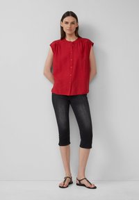 Czerwona bluzka z krótkim rękawem, z guzikami z przodu i marszczeniami, w połączeniu z czarnymi, ściętymi dżinsami i płaskimi sandałami.