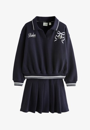 Giovane donna che indossa una felpa polo nera a maniche lunghe, gonna nera plissettata, calzini bianchi e scarpe da ginnastica bianche, in piedi davanti a uno sfondo chiaro e uniforme.