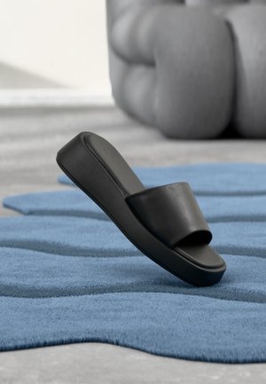 Μαύρο σανδάλι slide τοποθετημένο σε υφασμάτινο μπλε χαλάκι με μεγάλο γκρι μαλακό έπιπλο στο φόντο.