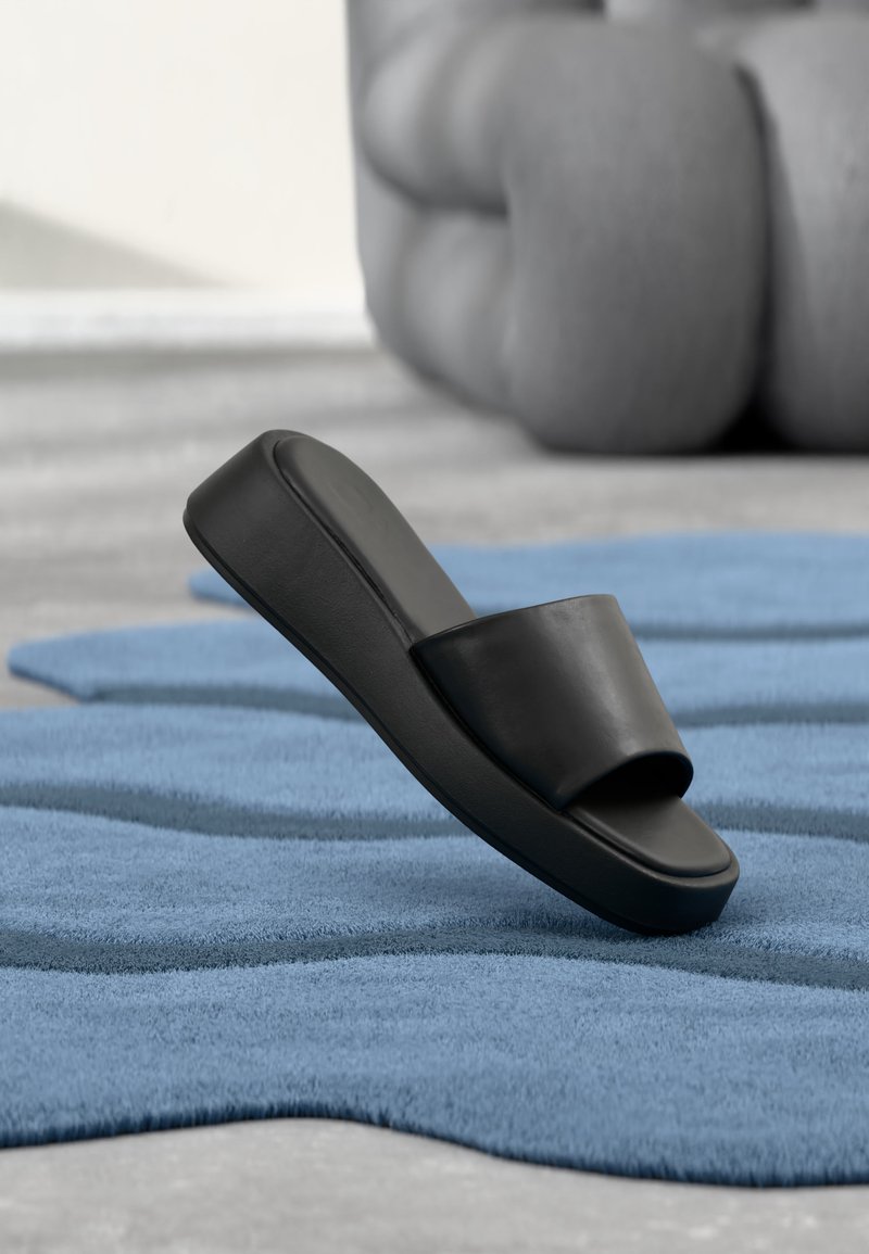 Μαύρο σανδάλι slide τοποθετημένο σε υφασμάτινο μπλε χαλάκι με μεγάλο γκρι μαλακό έπιπλο στο φόντο.