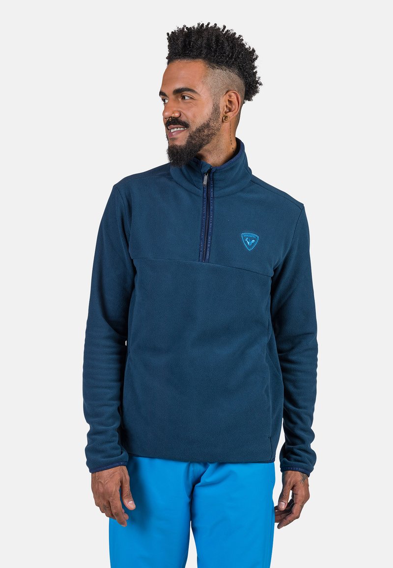 Mörkblå fleecepulloverjacka med hög krage, kvart zip-fram och en bröstficka med logotyp. Tyget har en mjuk textur.