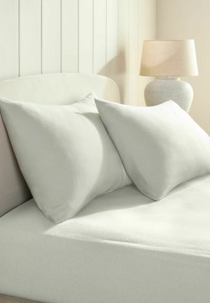 Zwei große weiße Kissen lehnen an einem weißen Kopfteil auf einem weißen Bett mit einer weißen Lampe auf einem Nachttisch.