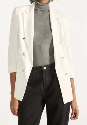 Person wearing a white double-breasted blazer over a gray turtleneck sweater and high-waisted black jeans, standing against a plain background.