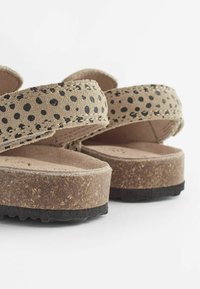 Beige Sandalen mit schwarzen Punkten, aus Wildleder, mit Kork-Zwischensohle und strukturierter schwarzer Gummi-Außensohle. Zwei gekreuzte Riemen auf der Oberseite.
