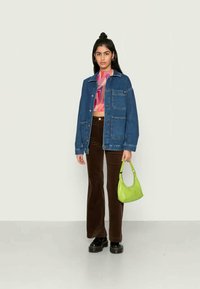 Jovem mulher vestindo uma jaqueta de denim, top colorido, calças castanhas, sapatos pretos, segurando uma bolsa verde limão contra um fundo simples.