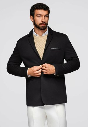 Schwarzer Blazer mit slim fit und Revers, ausgestattet mit einer Brusttasche. Getragen über einem cremfrohen Strickpullover und weißen Hosen. Strukturierter Stoff.