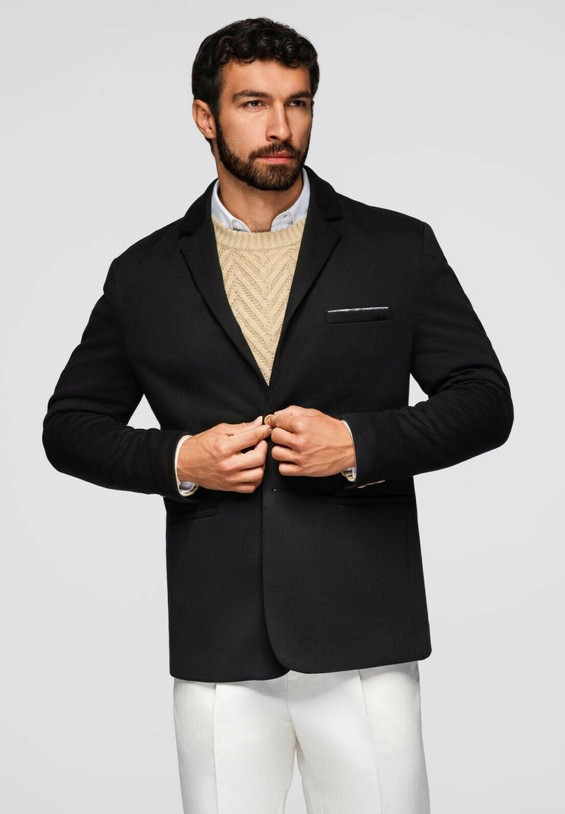 Schwarzer Blazer mit slim fit und Revers, ausgestattet mit einer Brusttasche. Getragen über einem cremfrohen Strickpullover und weißen Hosen. Strukturierter Stoff.