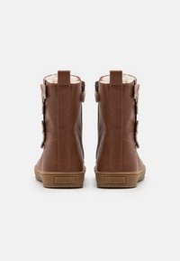 Hnědé kožené kotníkové boty s texturovaným povrchem, mají gumovou podrážku a zdobné boční přezky. Zadní pohled ukazuje výšku a designové detaily.
