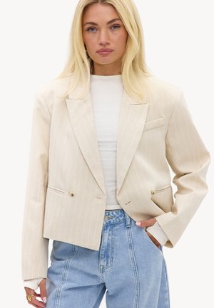 Femme blonde portant un blazer crème à fines rayures sur un top blanc et un jean bleu clair taille haute, une main dans la poche.
