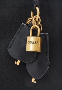 Gouden hangslot met de tekst "GUESS" en twee zwarte leren sleutelhoezen bevestigd aan gouden ringen op een zwarte, gestructureerde achtergrond.