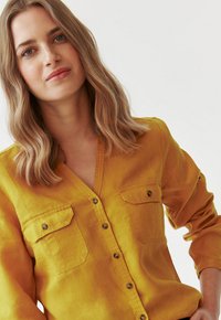 Mosterdgele linnen shirt met een V-hals, twee borstzakken en knoopdetails. Lange mouwen kunnen opgerold worden. Gladde textuur.
