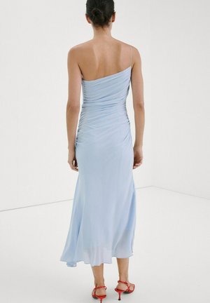 Mujer con cabello oscuro recogido en un moño, vestida con un vestido midi azul claro de un solo hombro y sandalias rojas de tacón, de pie en una habitación blanca mirando hacia otro lado.