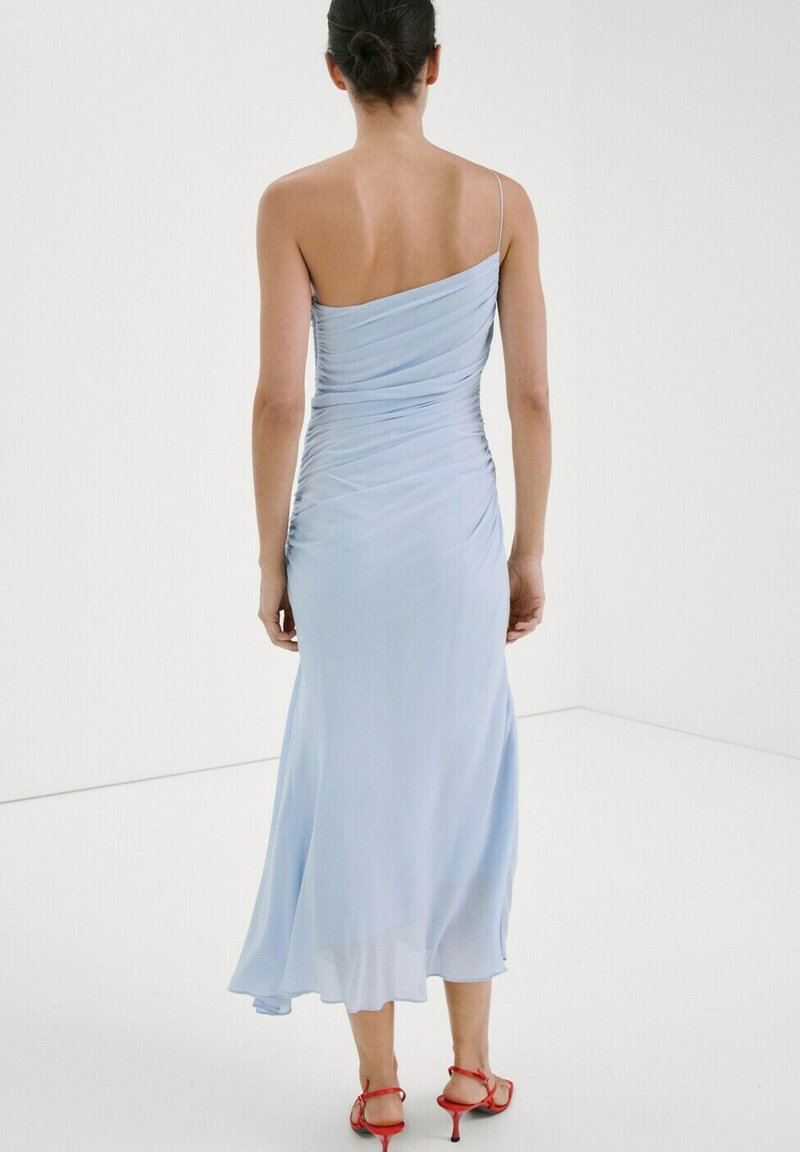 Femme aux cheveux noirs attachés en chignon, portant une robe midi bleu clair asymétrique et des sandales à talons rouges, debout dans une pièce blanche, tournée de dos.