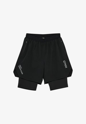 Jonge man staand, dragend een glanzend grijs jack met rits en capuchon, zwarte shorts met daaronder een legging, zwarte sokken en hardloopschoenen.