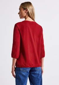 Donna con capelli biondi che indossa una camicia rossa a maniche lunghe e jeans blu, vista di spalle su uno sfondo bianco neutro.