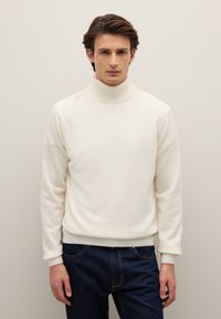 Cremefarbener Rollkragenpullover aus weichem Stoff mit geripptem Kragen und Bündchen, kombiniert mit dunkelblauen Jeans, glatte Textur.