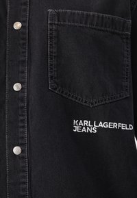 Juodas džinsinis marškinis su sidabro spalvos spaudėmis, krūtinės kišenė, ir išsiuvinėtas "KARL LAGERFELD JEANS" tekstas šone. Minkšta tekstūra.