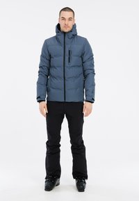 Dunkelblaue Herren-Puffjacke mit Reißverschluss, Kapuze und Vordertasche. Kombiniert mit schwarzen Skihosen und Stiefeln. Glatte Textur.
