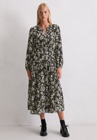 Czarna sukienka w kwiaty z dekoltem w kształcie litery V i długimi rękawami, zdobiona białymi i zielonymi wzorami kwiatowymi oraz warstwami materiału. Noszona z czarnymi butami.