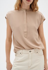 Beige blus med korta ärmar, hög krage och knapplist. Slät tyg med en subtil lyster. Bärs med ljusa jeans.