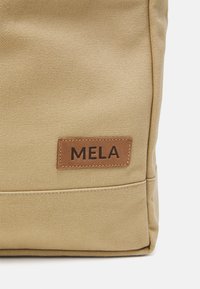 Khaki canvas tas met een rechthoekige vorm, textuurafwerking en een bruine leren label met "MELA" in reliëf.
