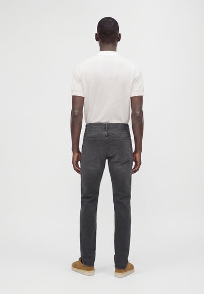 Homme debout de dos, portant une chemise blanche à manches courtes, un jean gris foncé et des chaussures marron sur un fond clair uni.