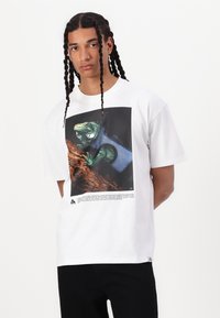 Tricou alb din bumbac, cu un grafic al unei iguane verzi pe un fundal întunecat, având text dedesubt și un design lejer, relaxat.