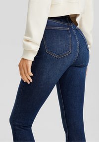 Tmavě modré skinny džíny s hladkou strukturou, středním pasem, designem s pěti kapsami a kontrastním šitím, předváděné vedle krémového topu.