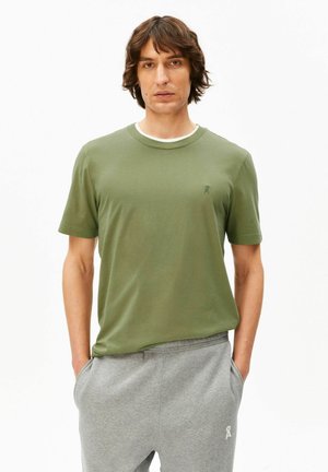 Homme portant un t-shirt vert olive et un pantalon de survêtement gris, les mains dans les poches, debout devant un fond blanc uni.