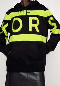 Sudadera negra con llamativas rayas amarillas y un gran lettering "KORS". Cuenta con capucha ajustable y bolsillo frontal, fabricada con material suave.