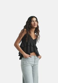 Svart lager av cami med volangdetaljer och prickar, matchad med ljusblå jeans. Tyget verkar lätt och strukturerat.