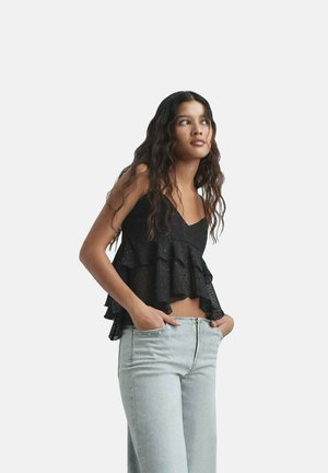 Čierna vrstvená top s volánikmi a bodkami, skombinovaný s svetlomodrými džínsami. Tkanina vyzerá ľahko a má textúrovaný povrch.