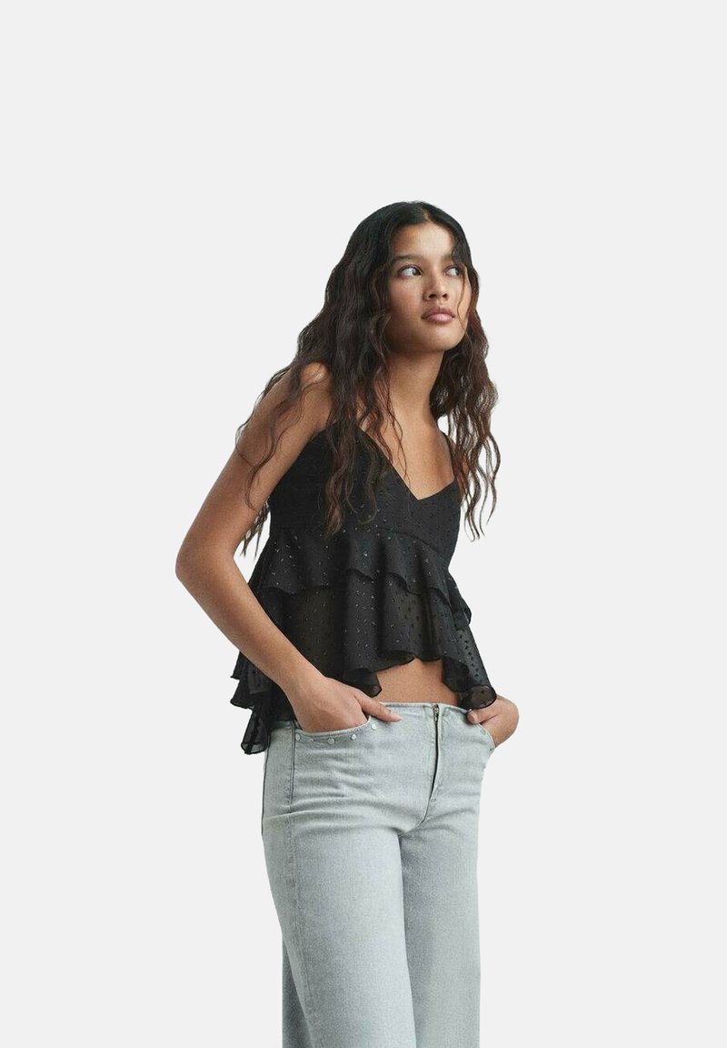 Svart lager av cami med volangdetaljer och prickar, matchad med ljusblå jeans. Tyget verkar lätt och strukturerat.